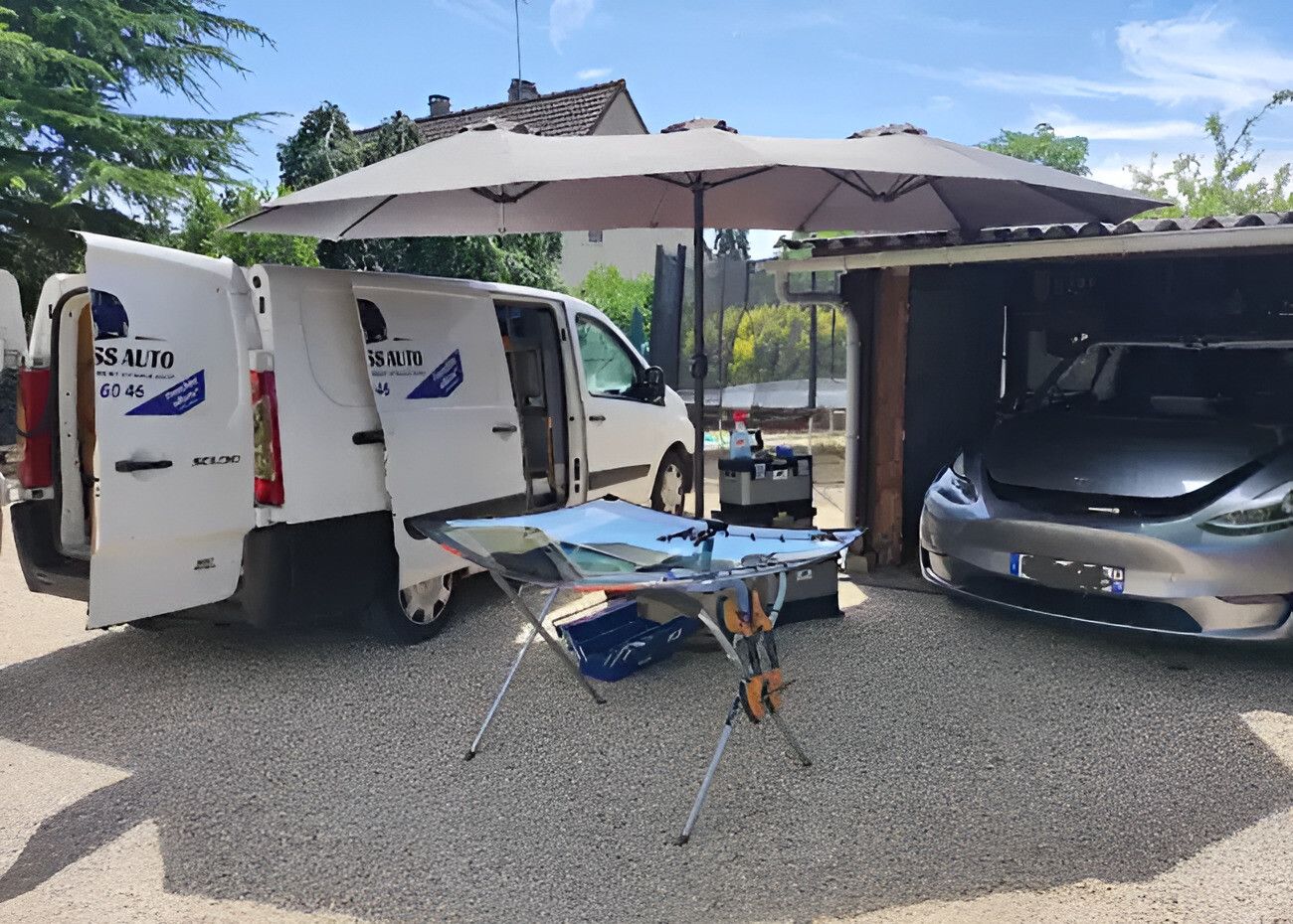 La camionnette Luso Glass Auto, portes ouvertes, et un pare-brise neuf sur son support sous un parasol, devant la voiture du client